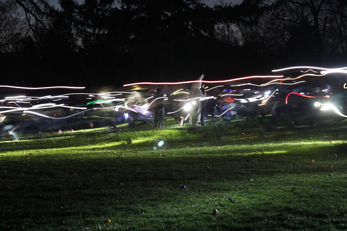 A time-lapsed photo of people running with flashlights: Multicolored streaks of light through a grassy area at night. There are dark silhouettes of trees in the background. Photo from Champaign Park District Facebook page.