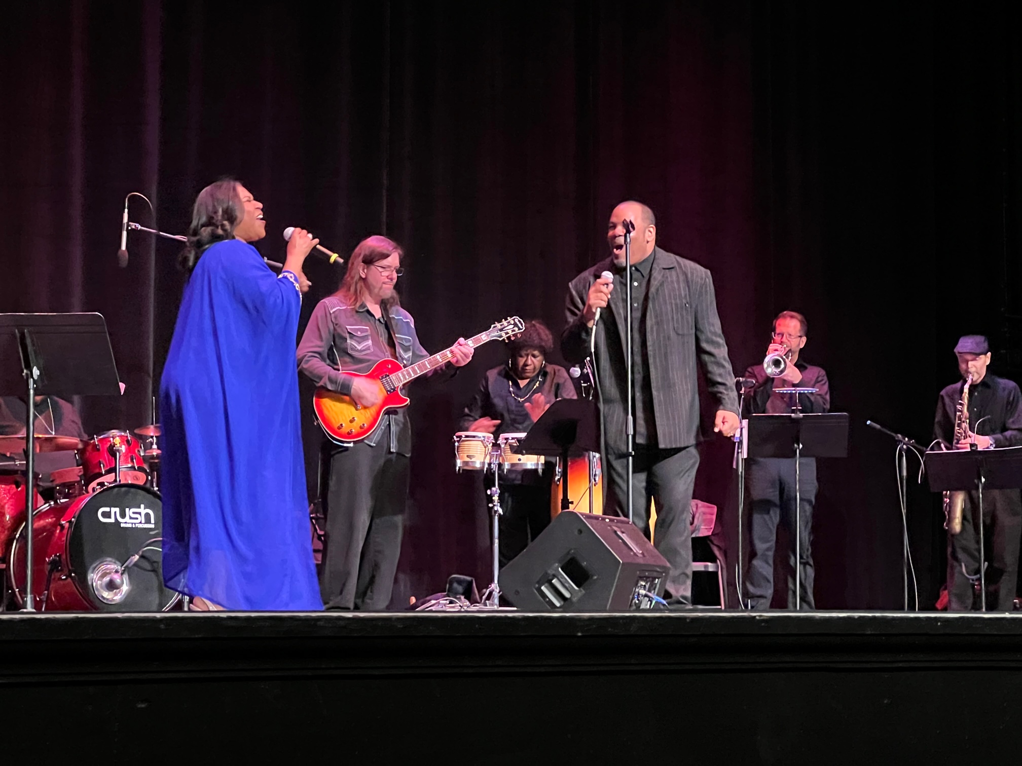 A Black woman in a flowing royal blue gown and a Black man in a black suit jacket, shirt, and pants, are facing each other on a stage, singing into microphone. In the background there is a white man with long hair playing a guitar. Also visible is a drum set, a Black woman playing bongos, and a white men playing a saxophone and trumpet. Photo by Julie McClure.