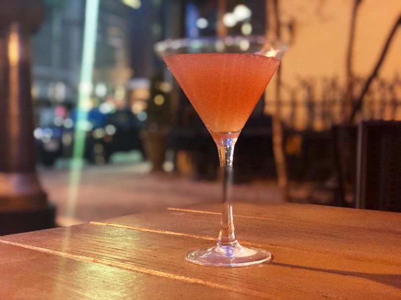 On a brown patio table outside of Cowboy Monkey, there is a pom splash in a martini glass at night. There is a light glare on the left side of the glass with blurred Downtown Champaign buildings lit in the background. Photo by Alyssa Buckley.