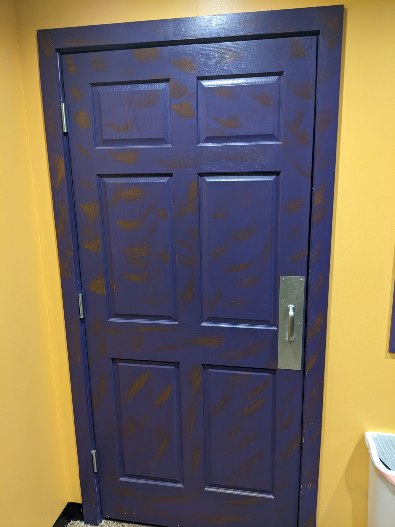A purple door with brown brush strokes scattered throughout. It has a gold handle. Photo by Tom Ackerman.