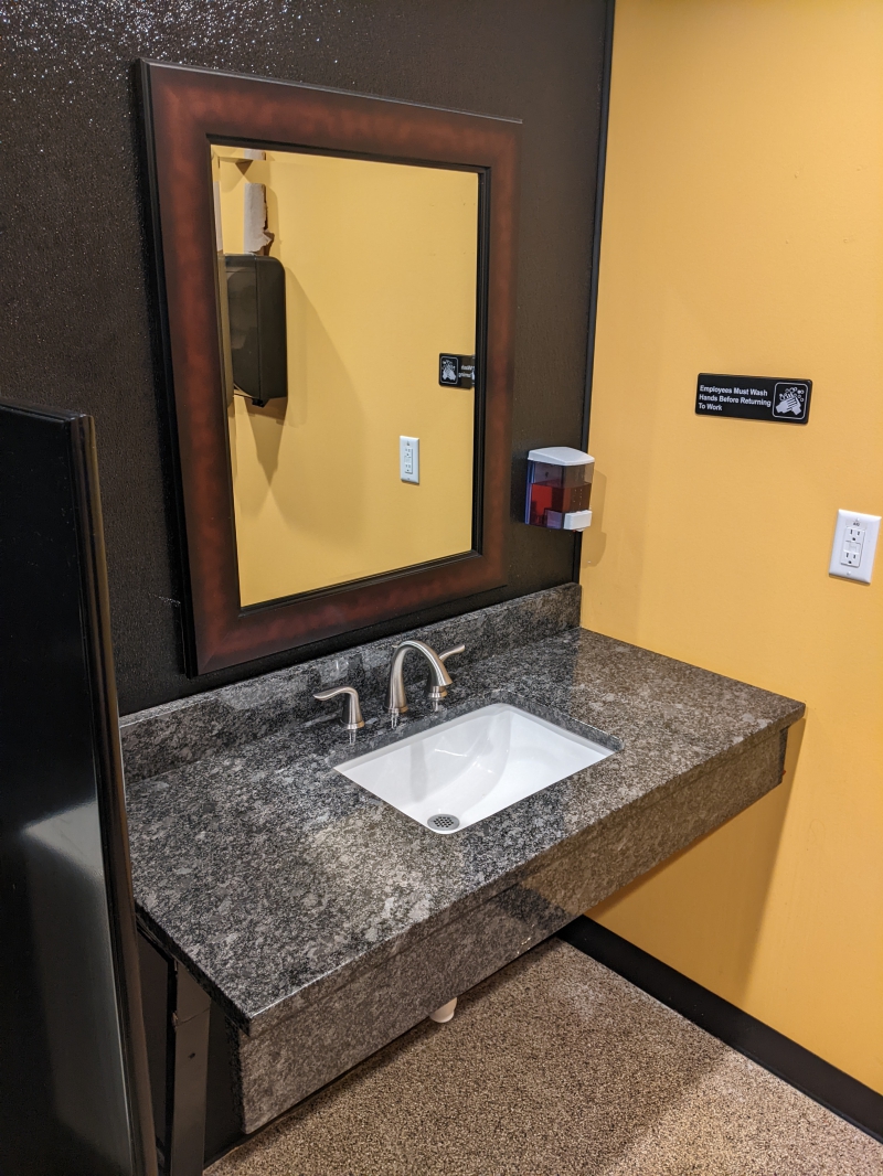 A dark gray marbled counter with a white rectangular sink in a restroom. A mirror hangs on a black wall behind the sink, and the adjacent wall is yellow. Photo by Tom Ackerman.