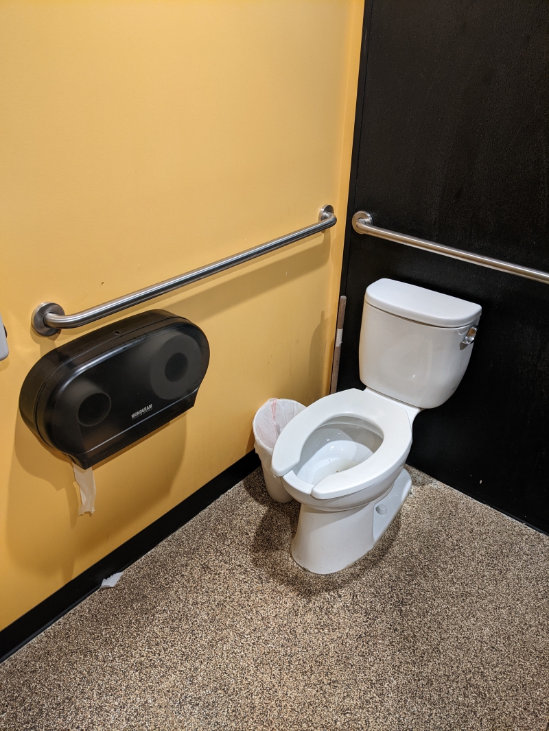 A white toilet in a stall with a yellow and black walls and a black toilet paper dispenser on the wall. The floor is black and white speckled. Photo by Tom Ackerman.