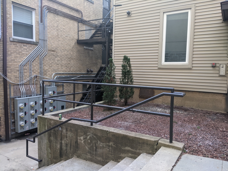 Concrete stairs with a black metal railing leading up to a concrete surrounded gravel patch with a couple of shrubs on the back side of a house. Photo by Tom Ackerman.v