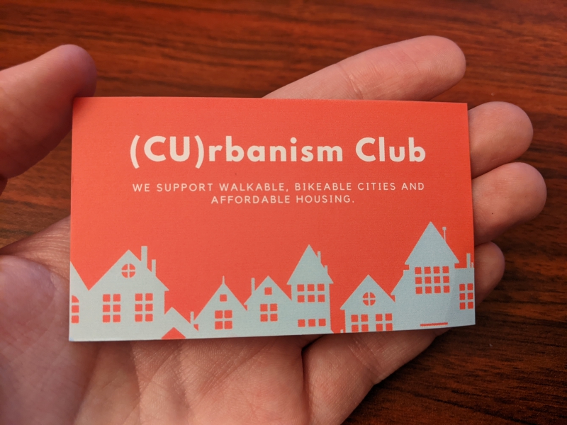 An orange business card with a white silhouette of a city along the bottom. It says (CU)rbanism Club in white lettering. It's resting in the palm of a hand. Photo by Tom Ackerman.