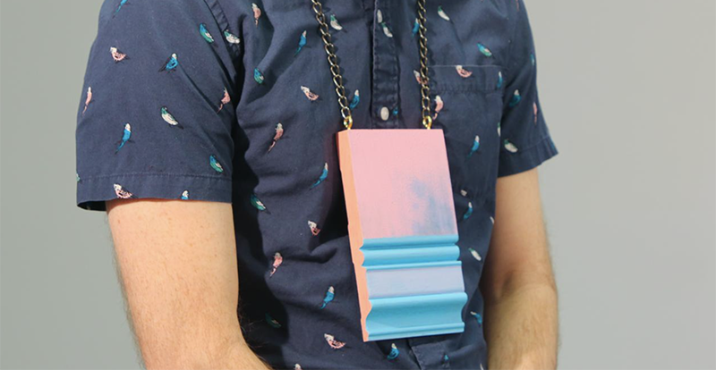 Photograph of man wearing a pink and large necklace made of pastel blue and pink painted baseboard material. 