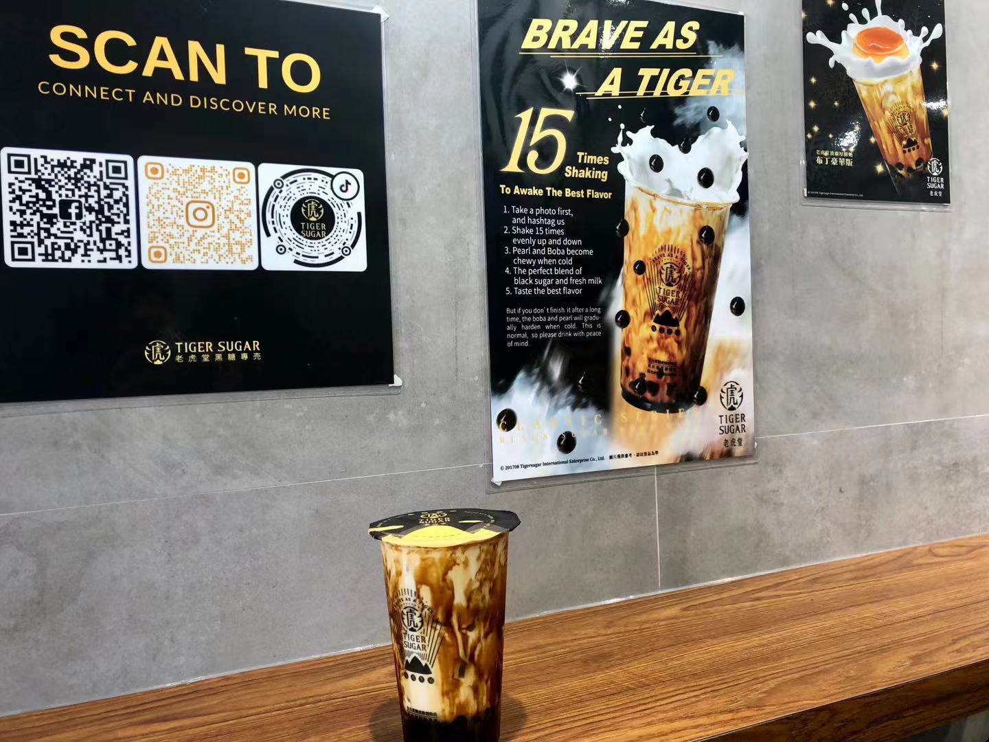 Inside Tiger Sugar, there is a boba drink on a wooden counter. Behind it, there are three boba posters on the gray wall. Photo by Xiaohui Zhang.