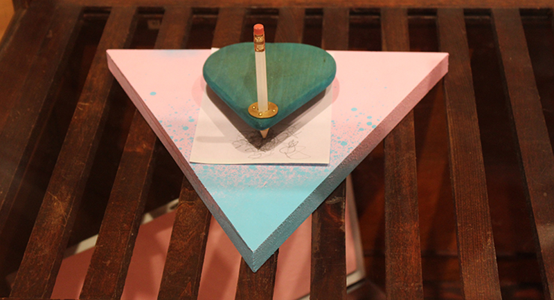 Wooden triangle painted pink and blue with a ouija board planchette held in place by a pencil.