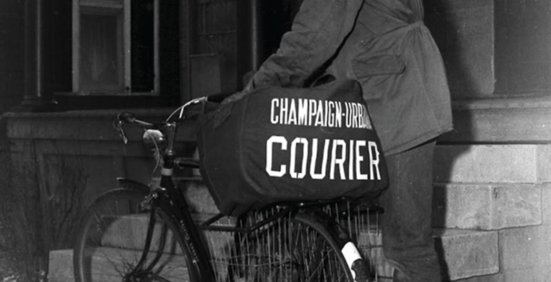 Black and white close up of newspaper delivery person riding a bike carrying a messenger bag with the name Champaign-Urbana Courier. 