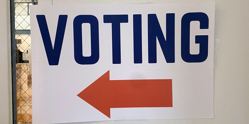 A white rectangular sign with blue block letters that say VOTING. There is an orange arrow under the word.