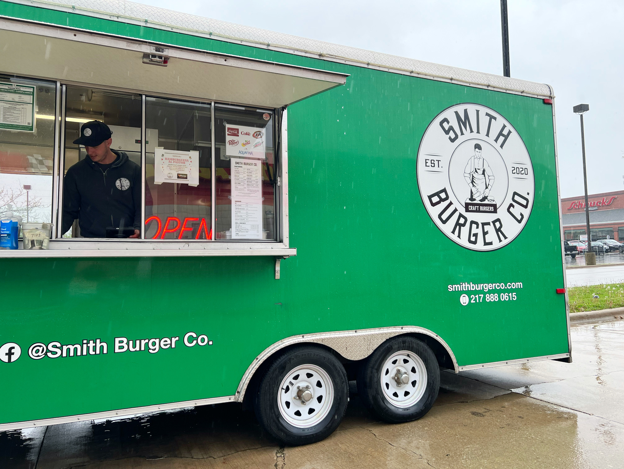 In a green food truck, there is a white man in a black hat and black hoodie. The green truck is Smith Burger Co, and the social media and company logo are printed on the side of the truck in white font. Photo by Alyssa Buckley.