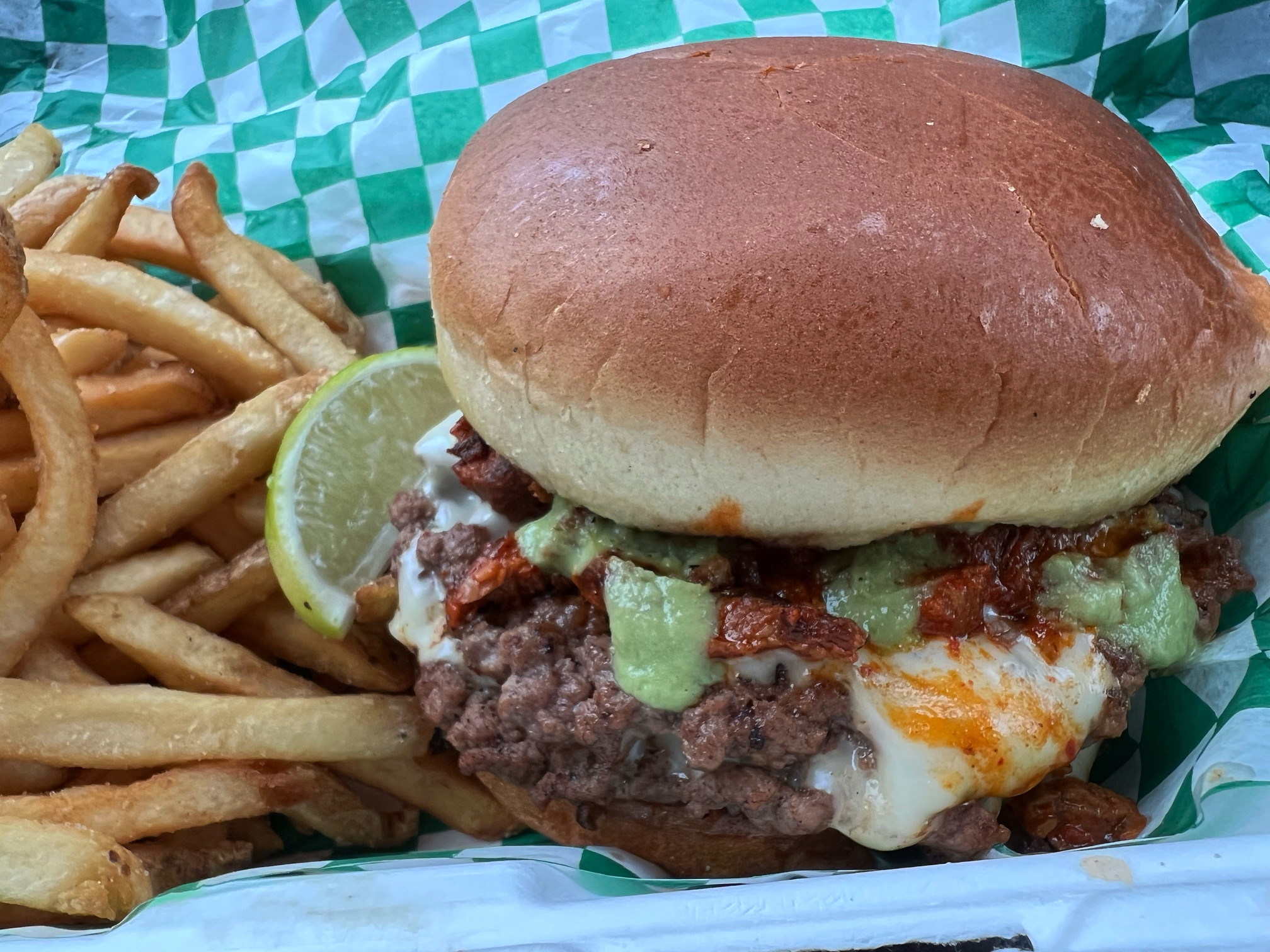 In a styrofoam container, there is a green and white checkered parchment paper with fries and a burger with guacamole salsa and melted white cheese. The burger is a collaboration of Smith Burger Co and Maize Mexican Grill for May 5th. Photo by Alyssa Buckley. 
