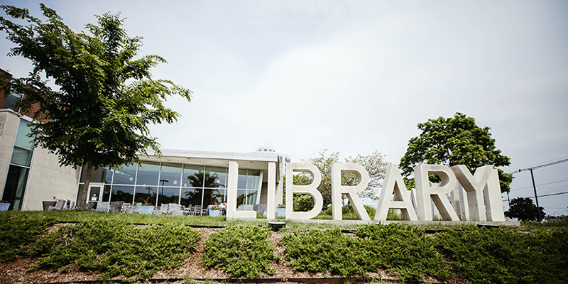 large white letters spelling out the word library sit on a small hill in front of a wall of windows. Clouds float above the building.