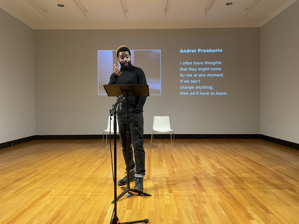 Brandon Burditt performs as part of Voices of the New Belarus at Krannert Art Musuem. He stands in front of a microphone. Behind him is a projection of the person whose words he is reading and the quote he is reading. Photo by Landon Sinclair.