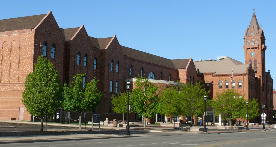 The Champaign County courthouse is a large, red brick building with a tower on the right side of the image. There are green trees in front of the building, along the sidewalk.