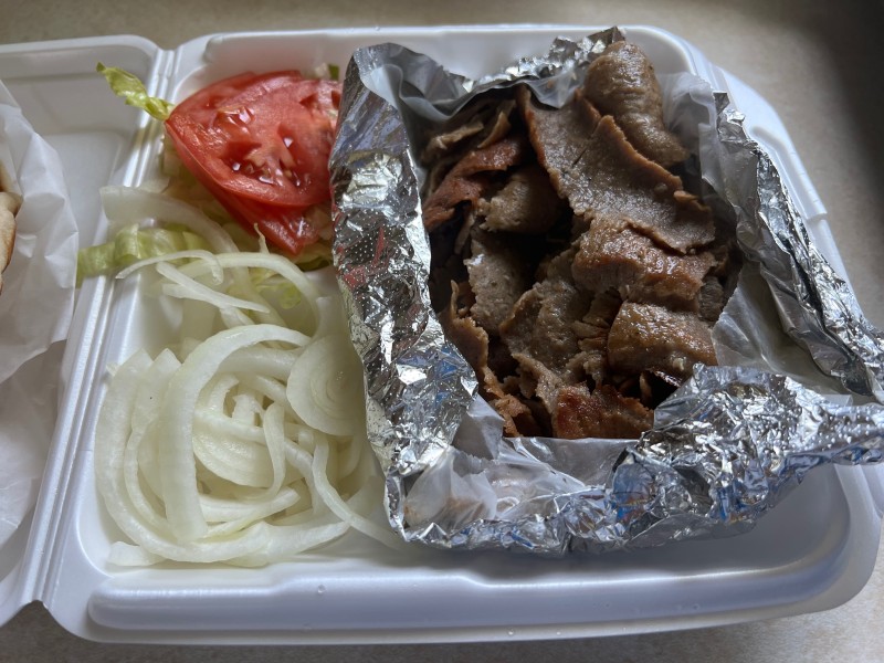 A white styrofoam take out container with thinly sliced gyro meat in a foil pouch on one side, and sliced white onions and red tomatoes on the other side. Photo by Julie McClure.