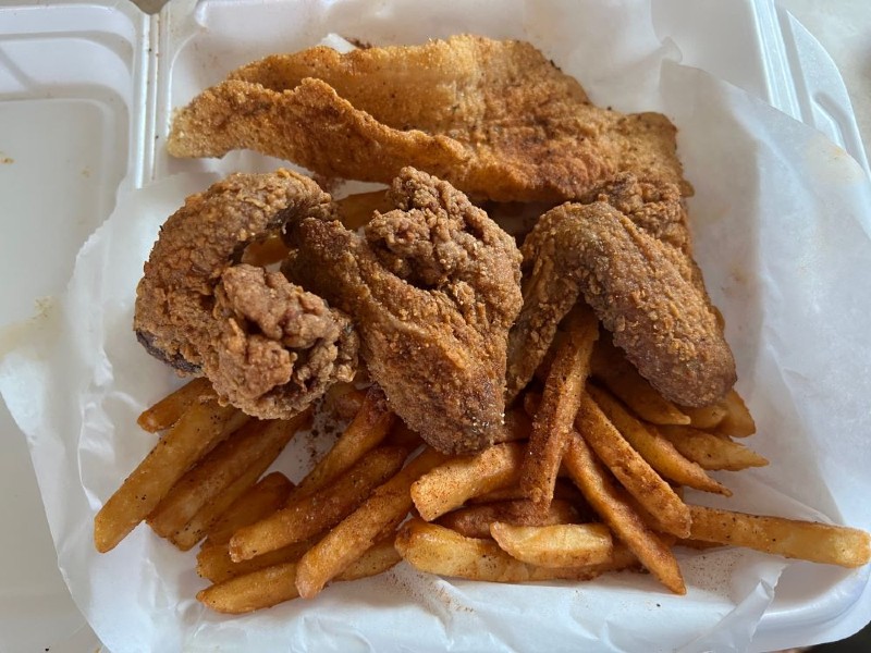 A white styrofoam take out container is filled with french fries, three pieces of breaded fried chicken chicken, and a long piece of breaded fried fish. Photo by Julie McClure.