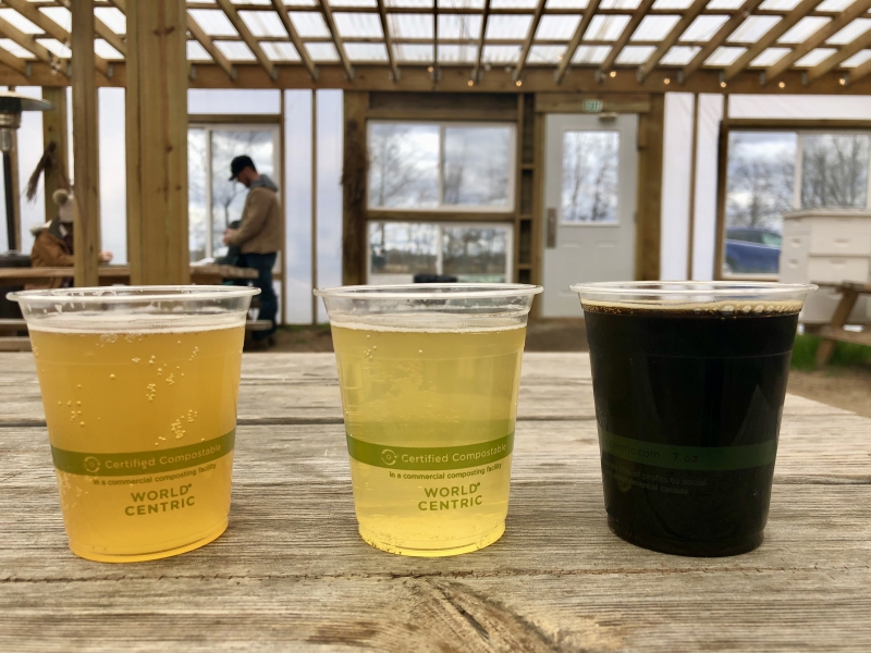 Three cold beers from Big Thorn Farm & Brewery sit on a weathered wooden bar. The drinks are in plastic cups. The two on the left are light colored and the one on the right is dark. Photo by Yobu (Arthur) Zou.