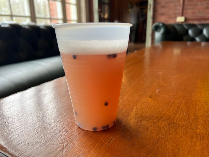 A clear plastic cup with a hazy orangish brown liquid sits on a wooden table. It has small dark berries resting on the bottom and floating at the top of the cup. Photo by Julie McClure.