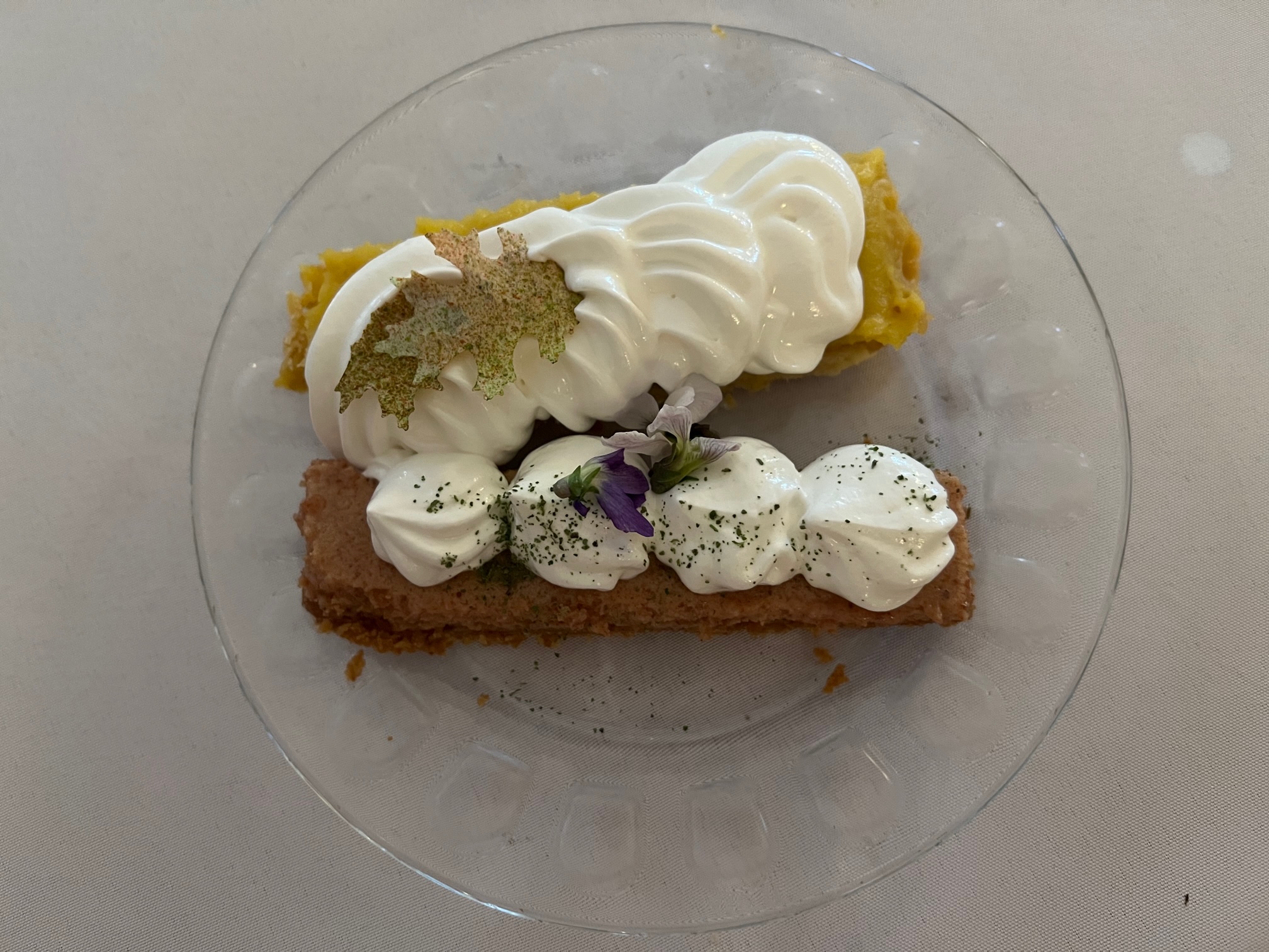 A small clear round plate on a white tablecloth. It is topped with two dessert bars, one yellow and one brown, and they are topped with swirled white cream. Photo by Julie McClure.