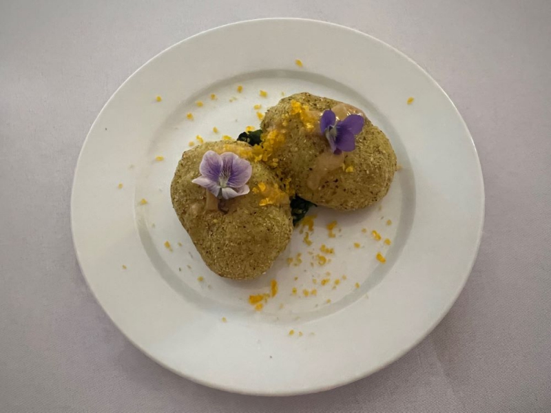 Two round potato croquettes on a small round white plate and white tablecloth. It is topped with yellow flakes and white and purple floral garnish. Photo by Julie McCLure.
