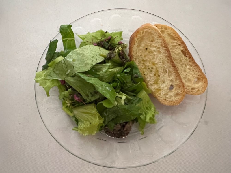 A round clear plate on a white tablecloth. It's topped with a leafy green salad and two slices of baguette on the side. Photo by Julie McClure.