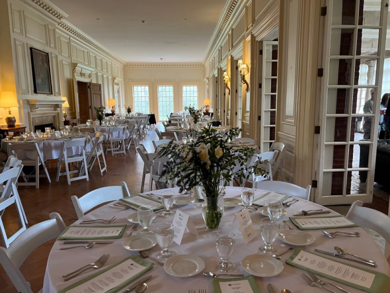 A long great hall with ornate ivory colored walls and three rectangular windows on the back wall. There are two rows of round tables covered in white tablecloths, and set for dinner. They have vases with white and yellow daffodils in them. Photo by Julie McClure.