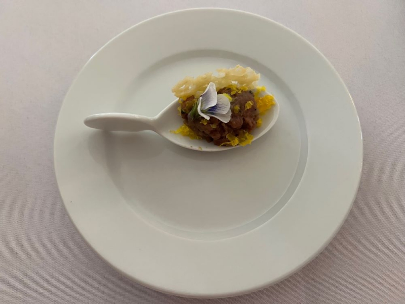 A round white plate on a white tablecloth holds a small white spoon filled with brown meat, a yellow crisp, and a white and purple bloom. Photo by Julie McClure.