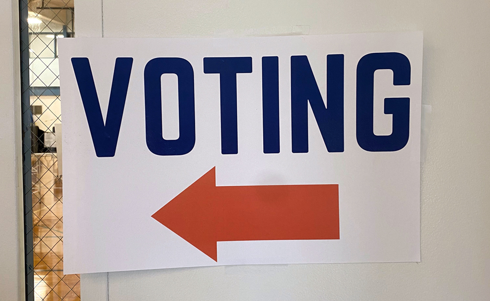A white sign that says VOTING in blue letters with an orange arrow pointing to the left. The sign is taped to a glass door. Photo by Jessica Hammie.