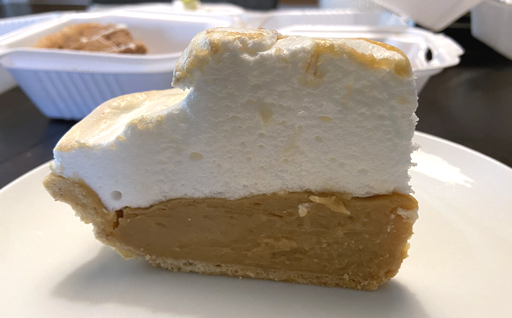 A slice of butterscotch meringue pie on a white plate on a table. The pie is a golden brown with a fluffy white meringue on top. Photo by Jessica Hammie. 