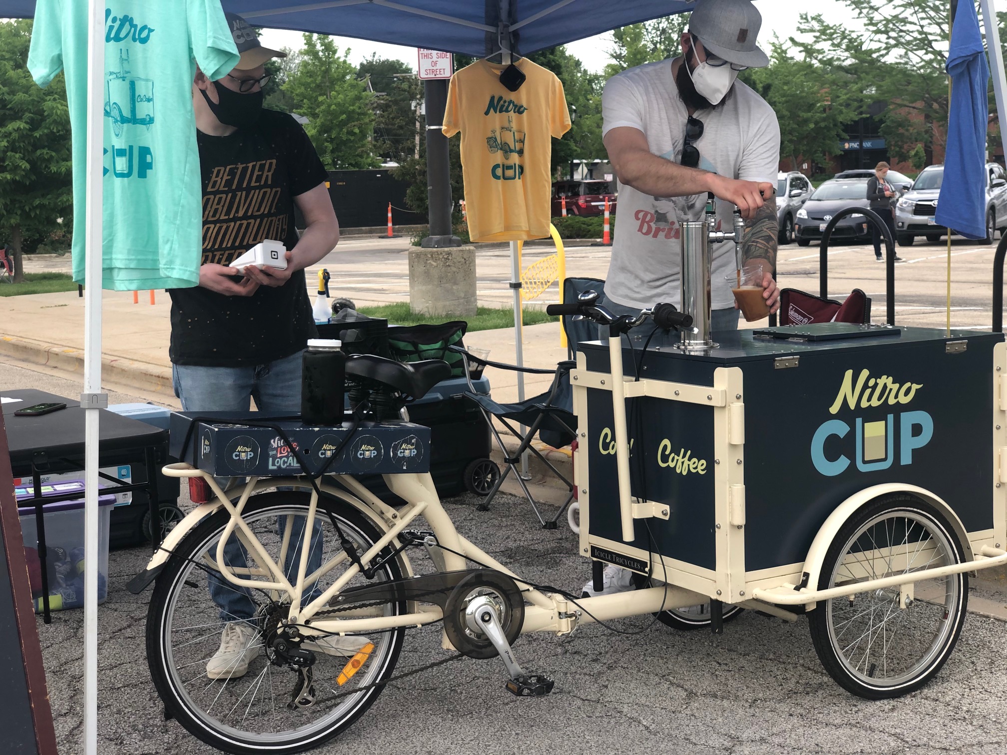At the Urbana Market at the Square, there is a coffeeshop on wheels and two white men working under a blue tent. One is pouring cold brew into a plastic cup.  Photo by Alyssa Buckley.
