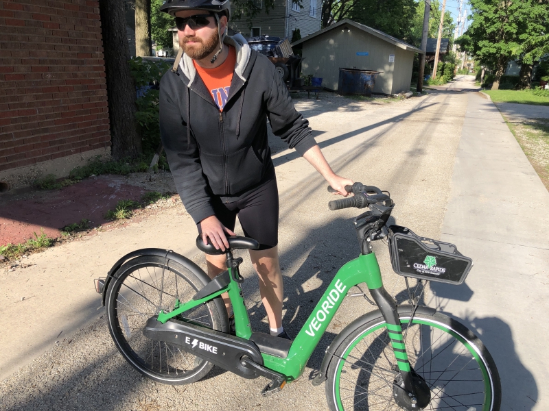 The writer is standing alongside a green electric bike. He's wearing a black bike helmet, black hoodie, black bike shorts, and sunglasses. Photo by Andrea Black.