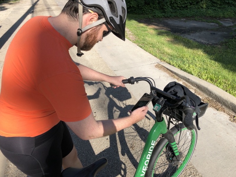 The writer is bent over a bike, taking a photo with his phone. He has on a bike helmet and an orange t shirt and black bike shorts. Photo by Andrea Black.
