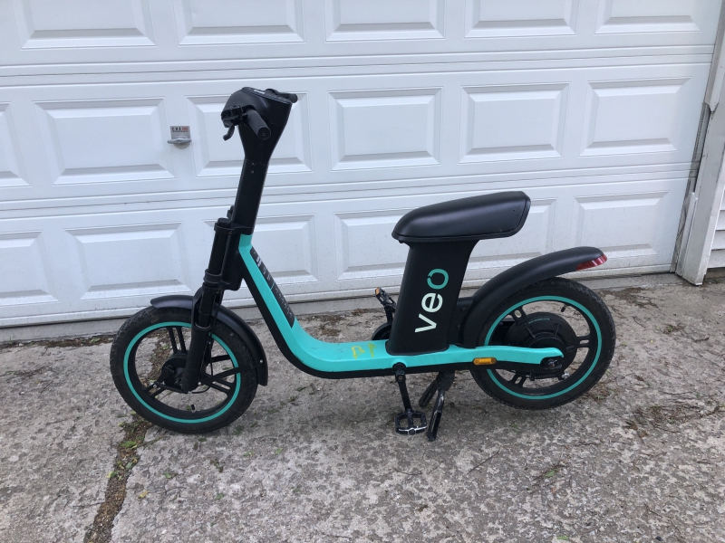 A black and turquoise motorized scooter with a seat. It sits in a driveway in front of a white garage door. Photo by Andrea Black.