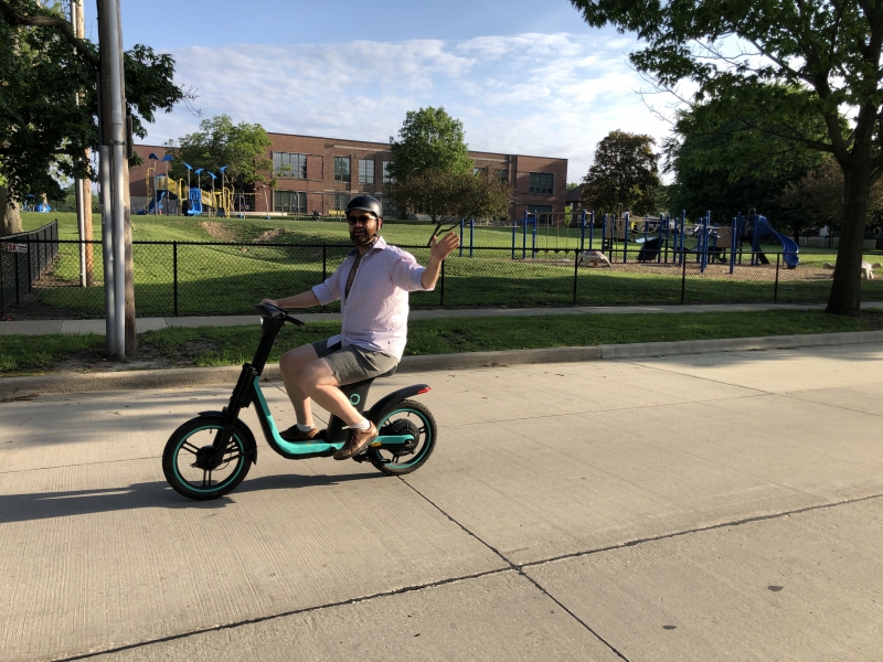 The writer is riding a scooter down the road, waving with one hand. There is a playground and school building in the background. Photo by Andrea Black.