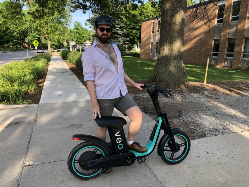 The writer is standing along side the motorized scooter with one foot propped on the base. He is wearing a white shirt that is partially unbuttoned and gray shorts. He is wearing sunglasses and a bike helmet. Photo by Andrea Black.