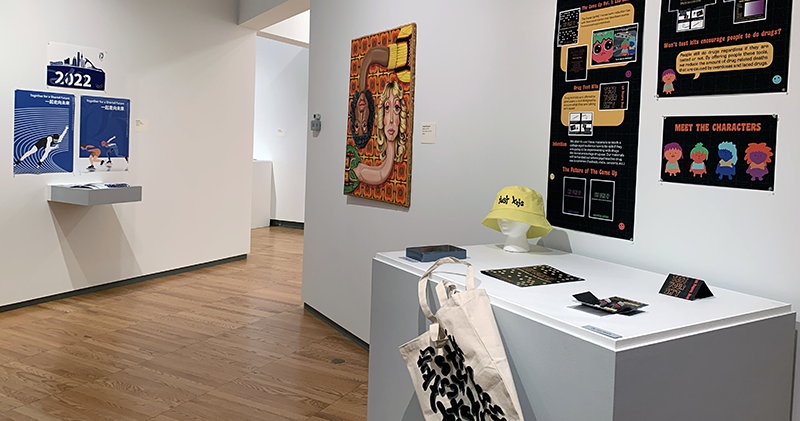 Wide view of gallery with paintings and posters on wall and design prototypes on pedestals. 