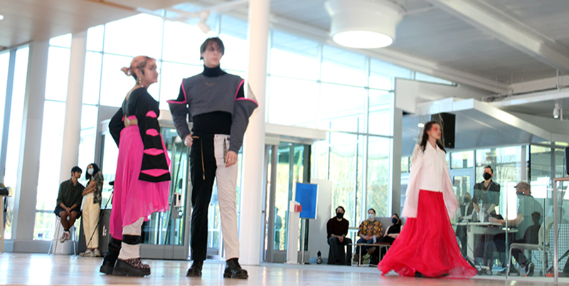 Female model in bright pink and black dress with cutouts with male model in black and white pants with black top and gray sleeveless vest and matching fingerless gloves with red trim. 