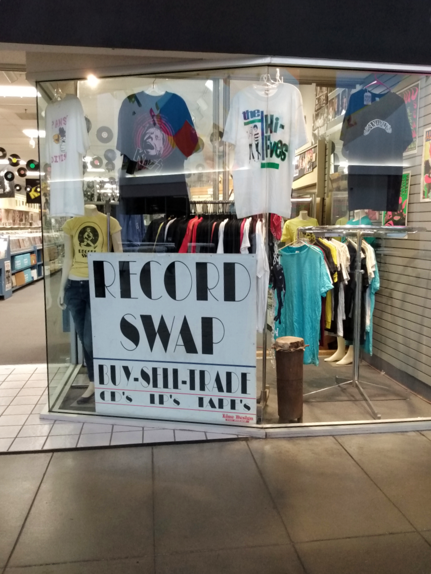 Record Swap's entrance in Lincoln Square Mall. A glass pane houses appreal with Record Swap's logo along with a sign for the store. Rows of record bins can be seen in the background.