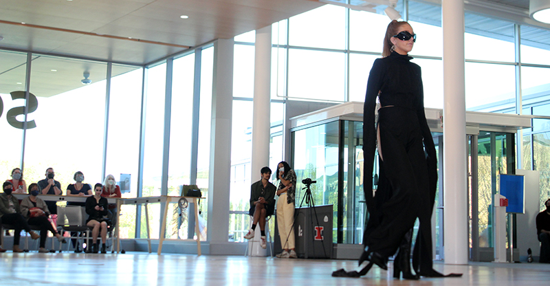 Female model in black high-necked body suit with long black strips on bottom hen and sleeves.wearing black sunglasses. 