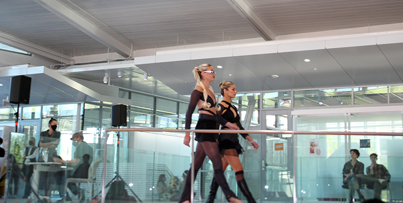 Sideview of two blonde female models in short black dresses with cutouts walking down ramp at Siebel Center.