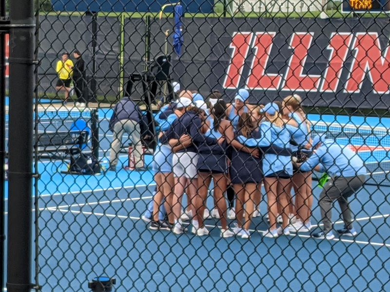 A view through a black chain link fence of a group of women's tennis players, wearing dark blue and light blue uniforms and white tennis shoes. They are in a huddle with their arms around each other. Photo by Sal Nudo.