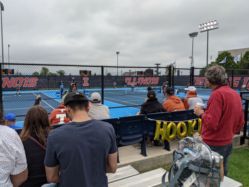 A view from the bleacher of a row of tennis courts. A black chain link fence separates the stands from the courts. There are people scattered throughout the stands, and large dark blue banners with Illinois in orange block letters on the opposite side of the courts. Photo by Sal Nudo.
