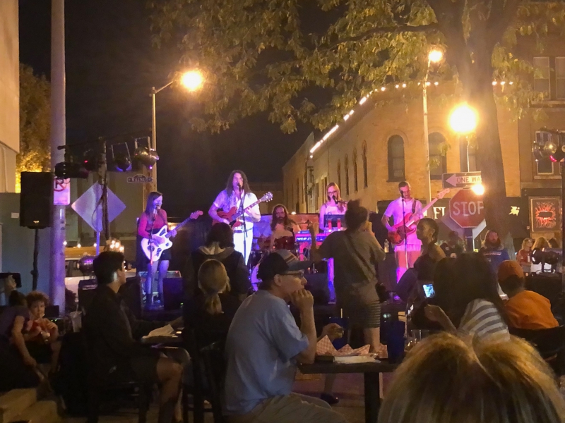 An outdoor concert at night. Several band members are on a stage in the distance. In the foreground are tables filled with people.