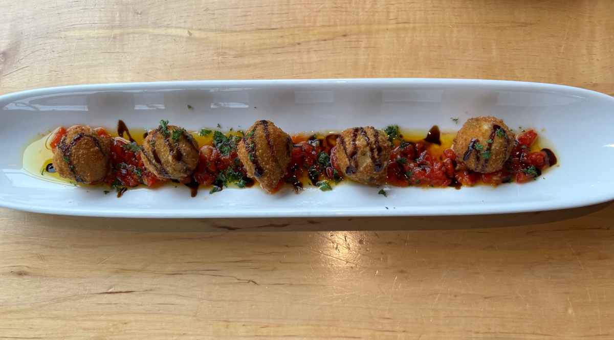 On a long white plate, there are five small arancini with a balsamic glaze on top of tomatoes and roasted red peppers. Photo by Alyssa Buckley.