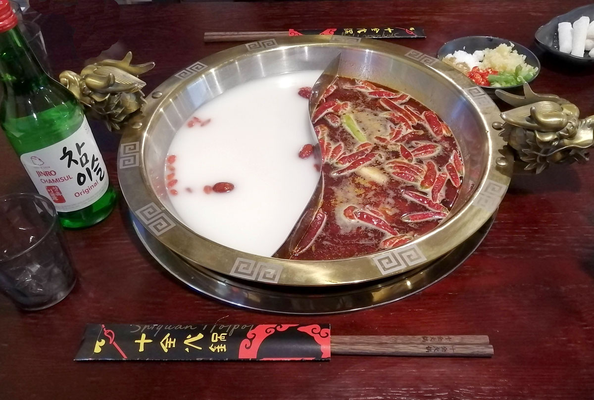 A split hot pot soup bowl is on a brown table with a bottle of soju beside it. Photo by Paul Young.