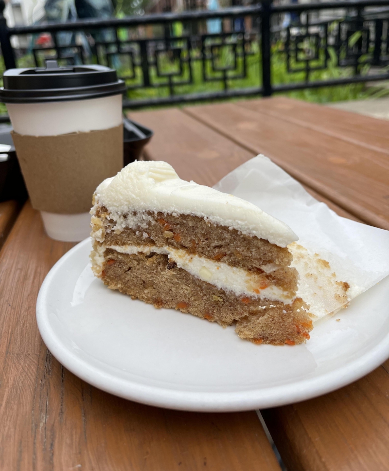 At Aroma Cafe on the outdoor patio, there is a slice of carrot cake dotted with nuts and frosted with white cream cheese frosting on a white circular plate. Behind the cake, there is a white paper to go coffee cup with a black plastic cover. 