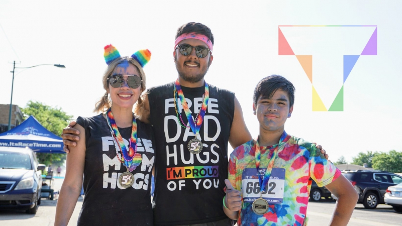 A woman, man, and teenager are standing arm and arm. They are all dressed up in rainbow accessories. The woman is wearing a shirt that says 