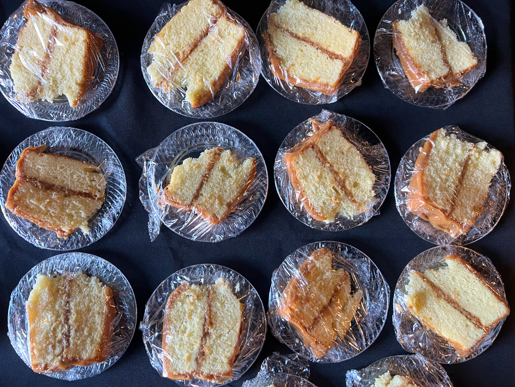 On a black table, there are lots of slices of vanilla cake with dark caramel frosting. All plates are clear plastic and there is plastic wrap around the slices. Photo by Alyssa Buckley.