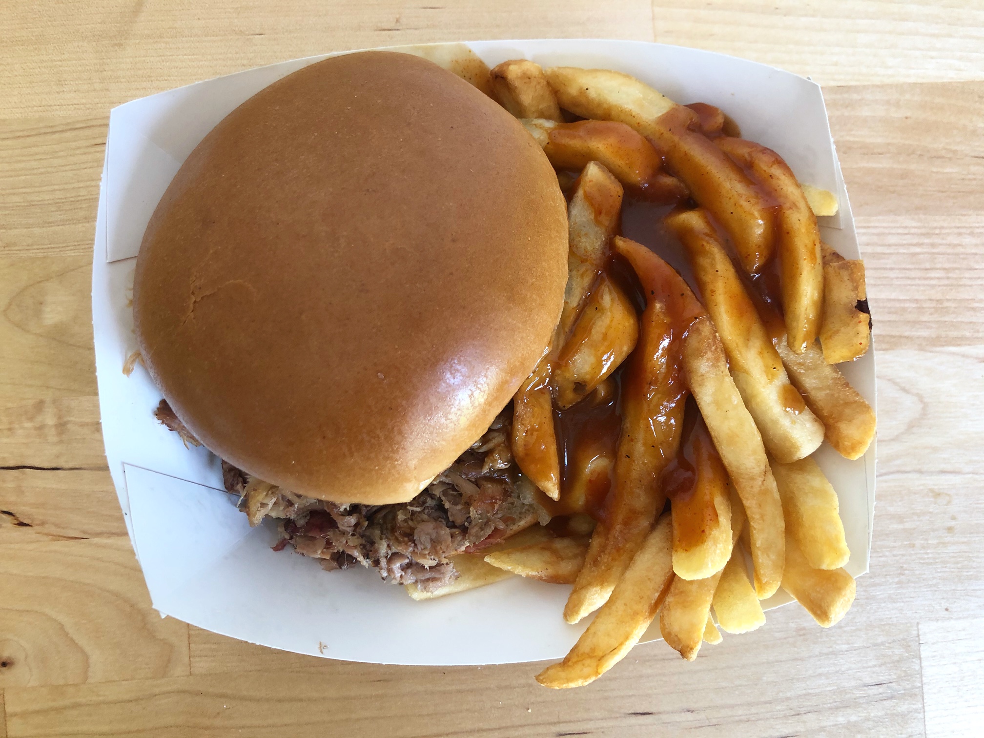 From above, a pulled pork sandwich in a perfect bun sits in a white paper boat with fries and a thick drizzle of barbeque sauce. Photo by Alyssa Buckley.