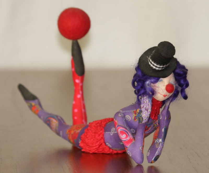 An image of one of Barbara Schoenoffâ€™s dolls, entitled, Tired Tumbler. The doll depicts a circus performer lying on her stomach, head raised and cradled on her hands, one leg raised, with a red ball balanced on the toes of her raised leg. The doll is resting on a smooth surface, a neutral background behind it.